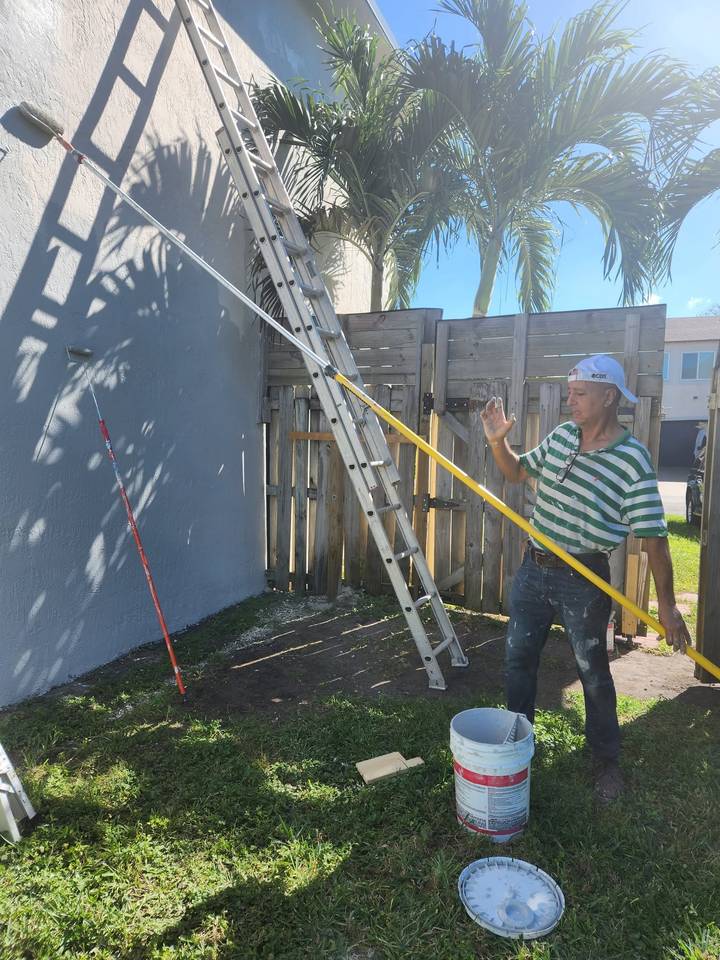 A1 Miami painter rolling fresh gray paint onto a stucco exterior wall with an extension pole, ladder and palm trees behind him